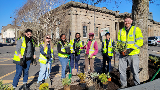 Scottish Water and Beautiful Perth Volunteers plant up the beds on Tay Street A group of seven people wearing high-visibility vests and gloves are planting greenery around a tree in an urban setting. They are standing on a paved area with a mix of potted plants, soil, and gardening tools. The background features a historic stone building with large windows and columns, as well as a clear blue sky. The group appears cheerful and engaged in their community gardening activity.