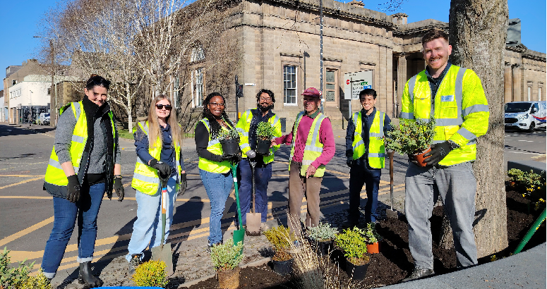 Scottish Water and Beautiful Perth Volunteers plant up the beds on Tay Street A group of seven people wearing high-visibility vests and gloves are planting greenery around a tree in an urban setting. They are standing on a paved area with a mix of potted plants, soil, and gardening tools. The background features a historic stone building with large windows and columns, as well as a clear blue sky.