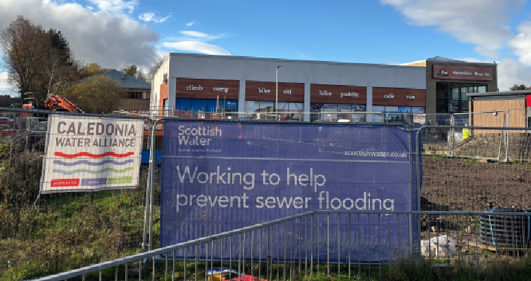 A construction site fenced off with Scottish Water signage reading “Working to help prevent sewer flooding.” Another banner for the Caledonia Water Alliance hangs on the fence. Behind the site is a retail building with signs for outdoor and sports shops. The sky is partly cloudy with trees visible to the left.