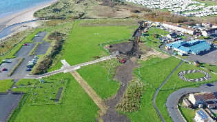 Aerial view of a coastal construction site showing excavation work on a grassy area near the beach. Machinery and vehicles are visible along a long trench, with nearby car parks, houses, and a caravan park in the background.