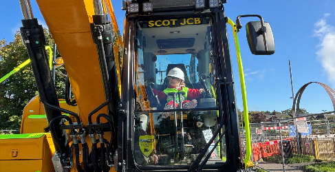 A child sits in a digger on a construction site. The sky in the background is blue.