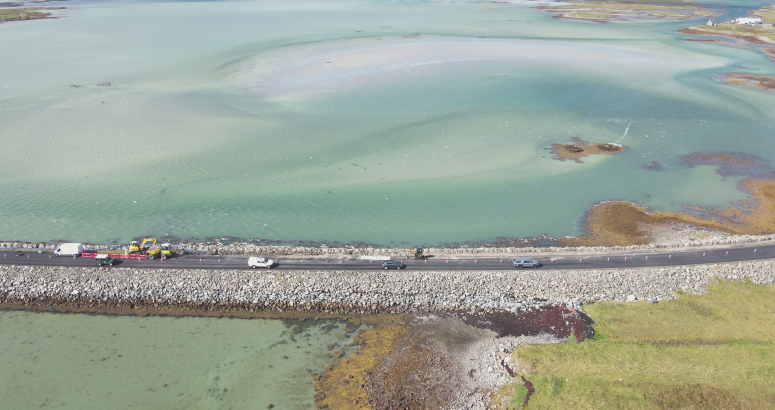 Benbecula Drone Footage Aerial view of a coastal road with cars and contractor vehicles near a large, shallow turquoise and green bay with visible aquatic plants and sand patches.