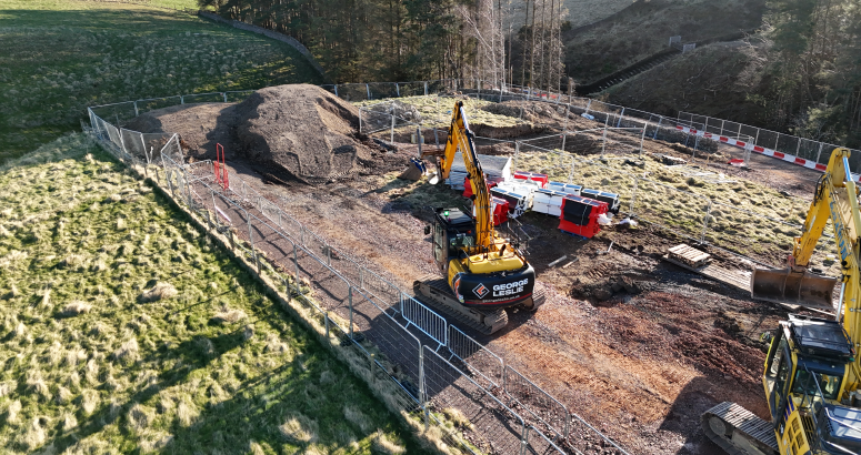 Aerial view of a construction site with several excavators, fenced-off areas, and construction materials scattered around, set in a grassy, slightly forested area.