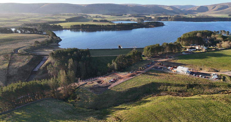 An aerial view of a construction site near a large body of open water with surrounding green landscape.