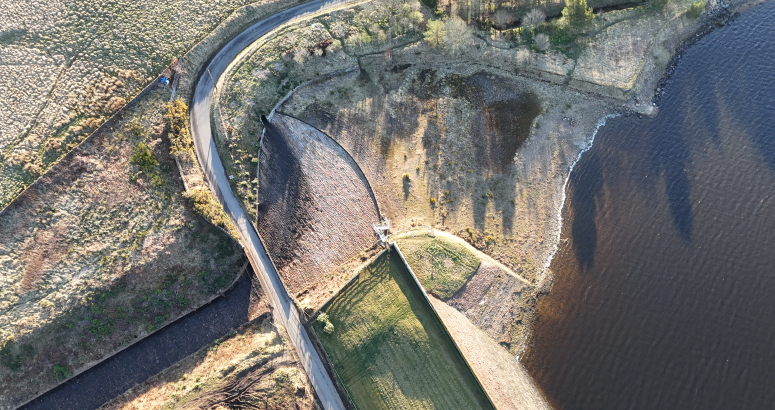 Aerial view of a curving road alongside a dam, with water on one side and a rugged landscape on the other.
