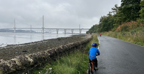 Thomas and Murray cycling with views of the Forth Bridges in the background Two children in bright clothing cycling on tarmac road with bridges in the background