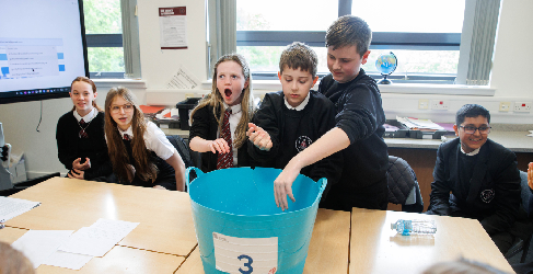 Students Take on Cold Water Challenge Students at Trinity High School gather around a large blue tub during a classroom challenge; one student reaches into the tub while others look on with excitement and surprise.