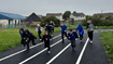 Children running along a running track. Grass surrounds the track and buildings can be seen in the background. The sky is grey.