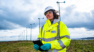 Economic Impact Report Scottish Water employee in PPE holding a tablet in front of a wind farm