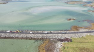 South Ford Causeway between the island of Benbecula and South Uist Aerial view of a causeway crossing a shallow, turquoise bay with scattered vehicles parked along the sides.