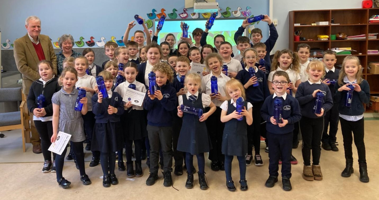 Scottish Water visits Benbecula school to deliver water-cycle talk Group of schoolchildren proudly displaying Scottish Water reusable water bottles in a classroom, with an adult supervising in the background.