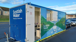 A mobile customer unit branded with Scottish Water logo and with an open white door. It's sat outside on a grey road with pavement in foreground and blue skies in background.