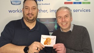 Two individuals standing in front of a Scottish Water banner, one holding a Thank You card from a customer with an image of a highland cow on it.