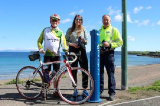 Male Scottish Water employee, female board member of Thurso Community Development Trust, and male from Caithness Cycling Club pose on sunny day beside newly-installed Top Up Tap Locals celebrating Top Up Tap launch