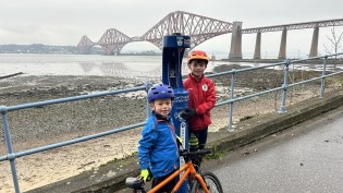 Thomas and Murray beside South Queensferry Top Up Tap Two young children pose beside bright blue water refill fountain with several large bridges in the background
