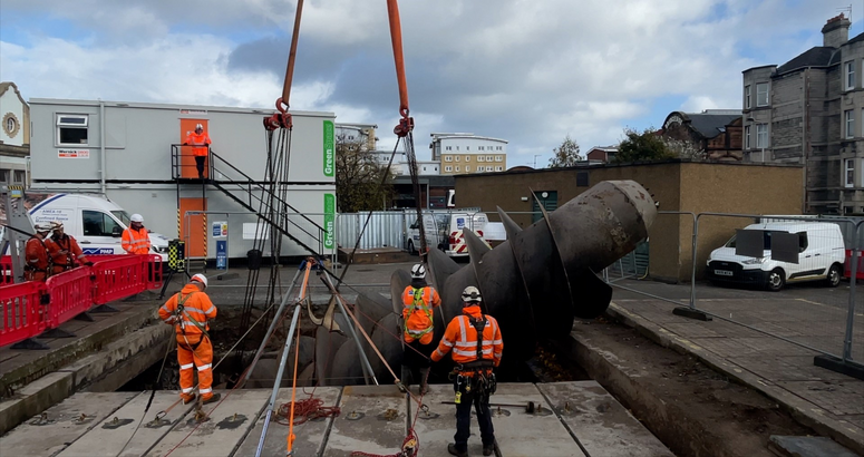 A group of people wearing hi-vis and PPE are standing beside a 9 tonne metal screw being lifted out of the ground. There is plastic fencing to mark the boundary and site portacabins in the background, with a white vehicle and properties to the right hand side.