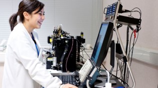 Scientist in lab coat using a laptop with advanced laboratory equipment in the background.