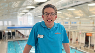 Scott Quin, a swim teacher at Edinburgh Leisure, standing in front of a swimming pool inside a brightly lit leisure centre.